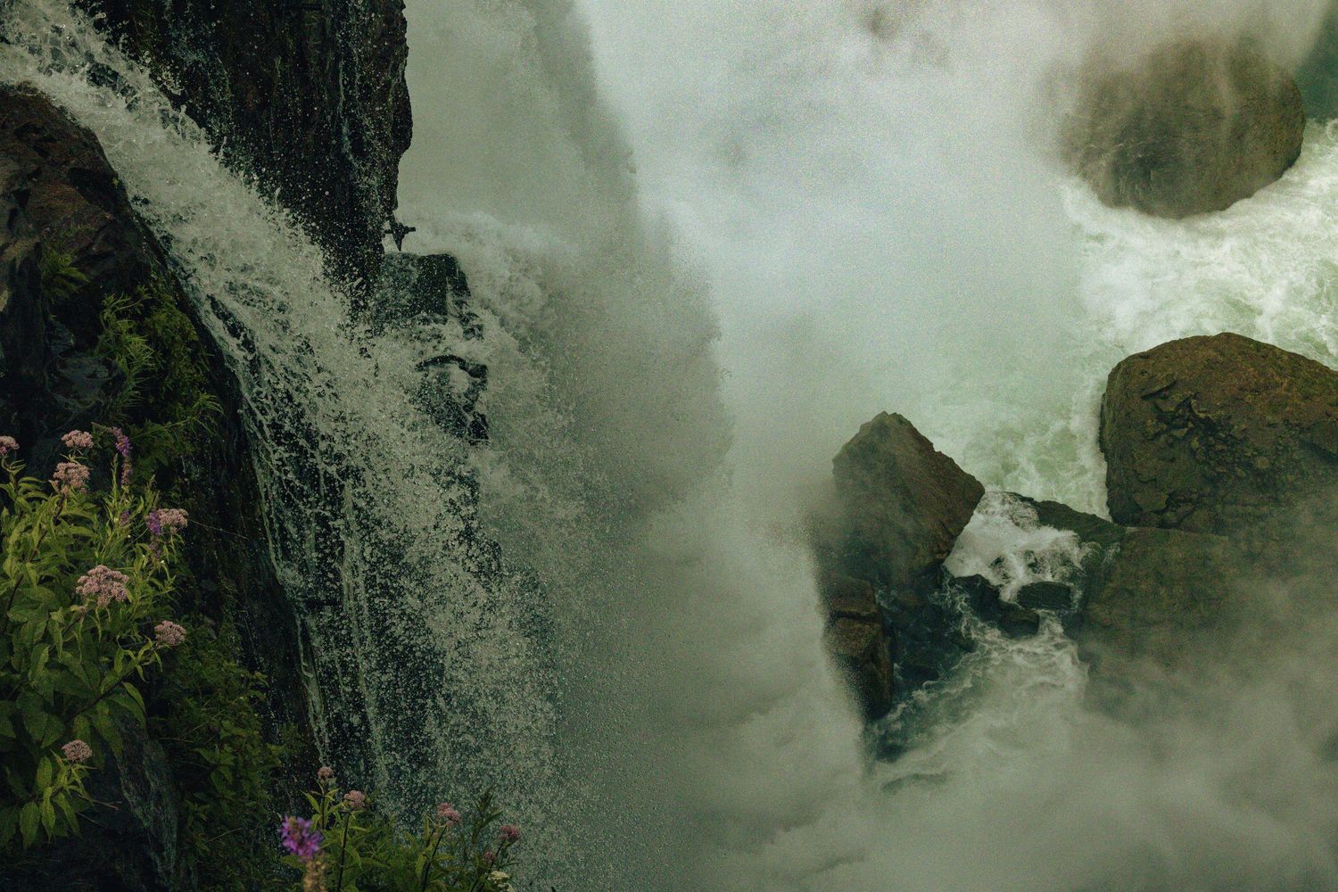 Powerful waterfall cascading over rocky cliffs with mist and pink wildflowers growing on the rugged stone surface.