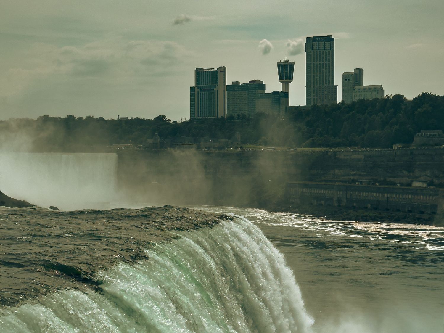 Niagara Falls rushing over the edge with misty spray and Canadian city skyline in the background.
