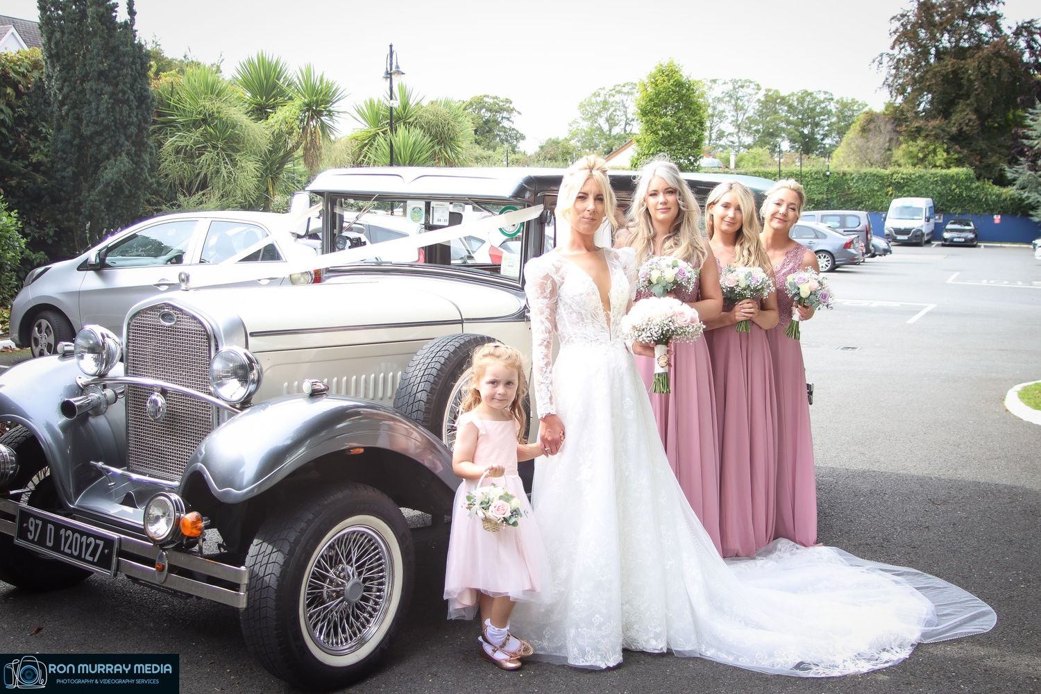 Wedding party poses with a classic vintage car featuring bridesmaids in dusty rose dresses.