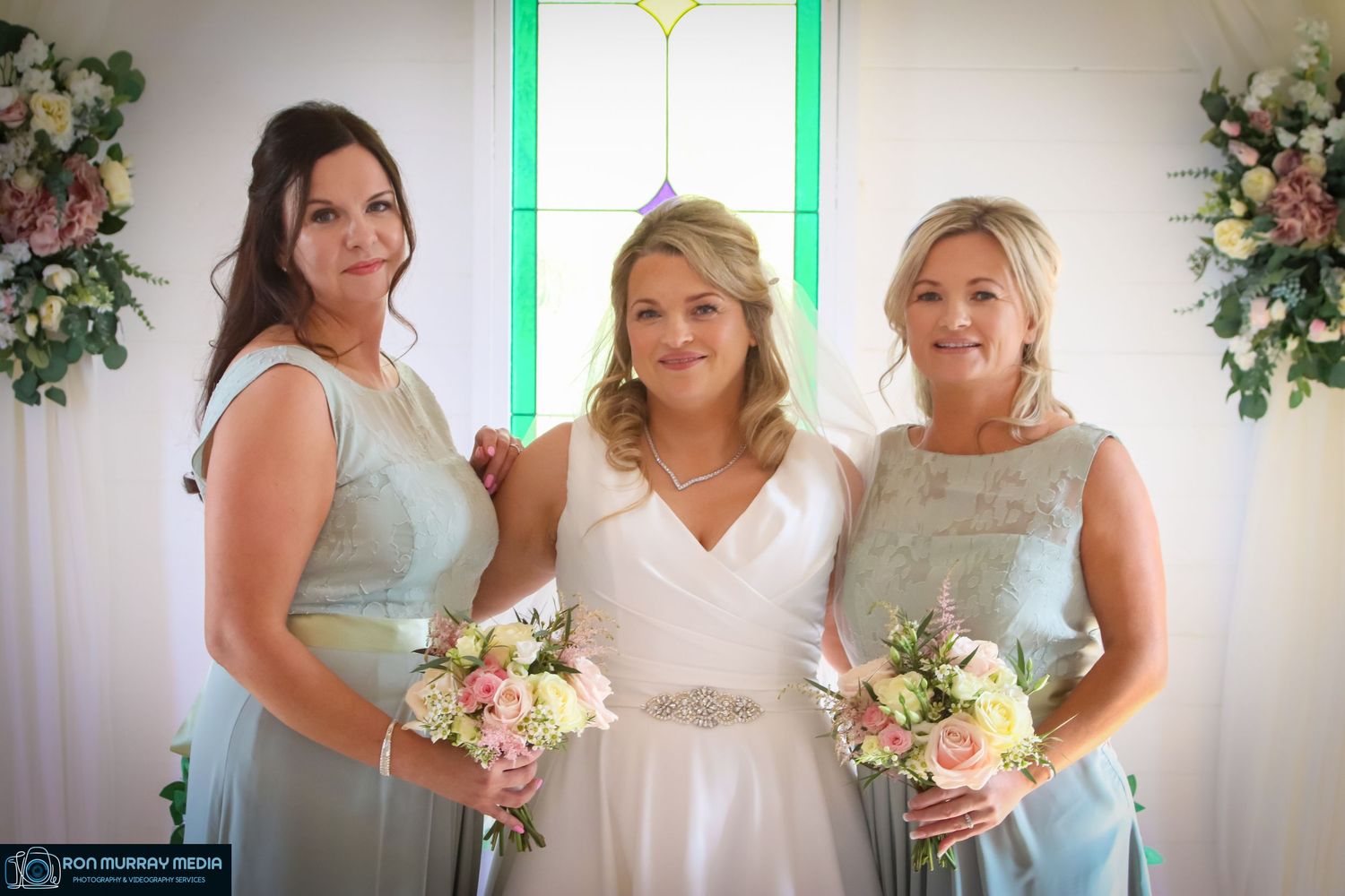 Three women in formal attire pose with matching bouquets against a floral backdrop.