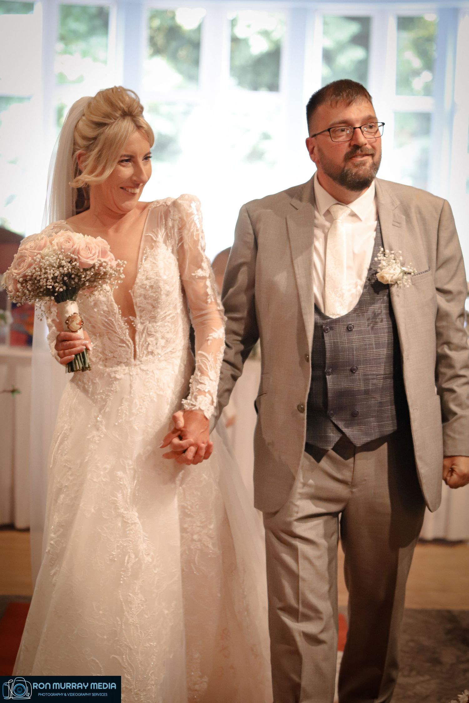 A couple walks hand-in-hand in formal wedding attire through a bright sunlit room.