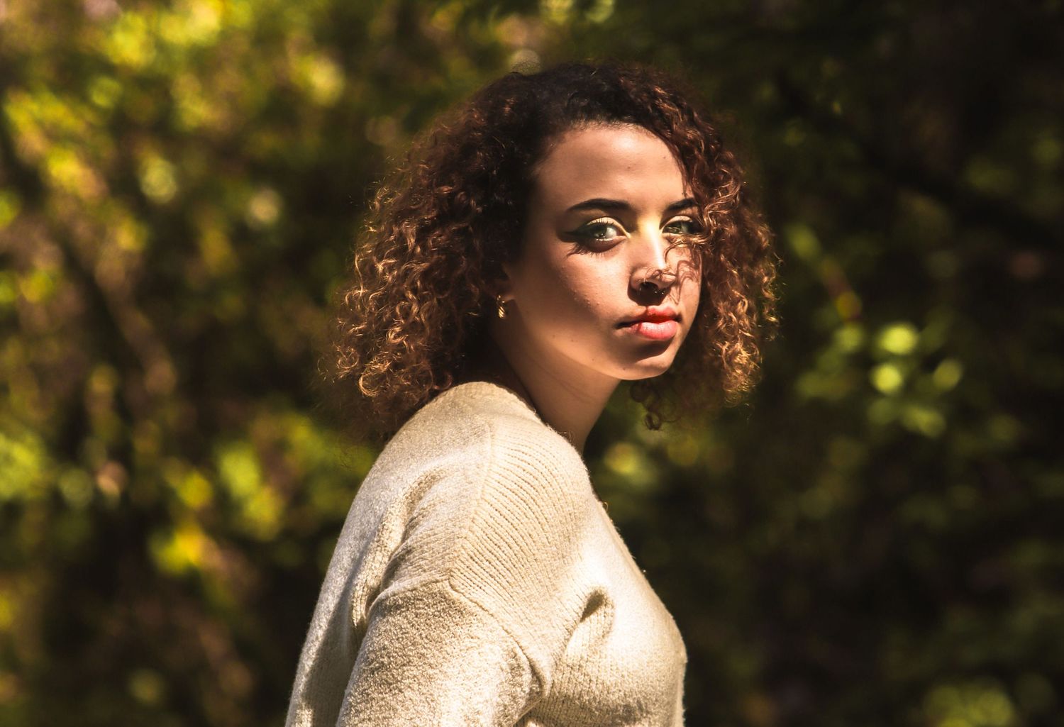 A person with curly hair poses outdoors in warm sunlight wearing a cream-colored sweater.