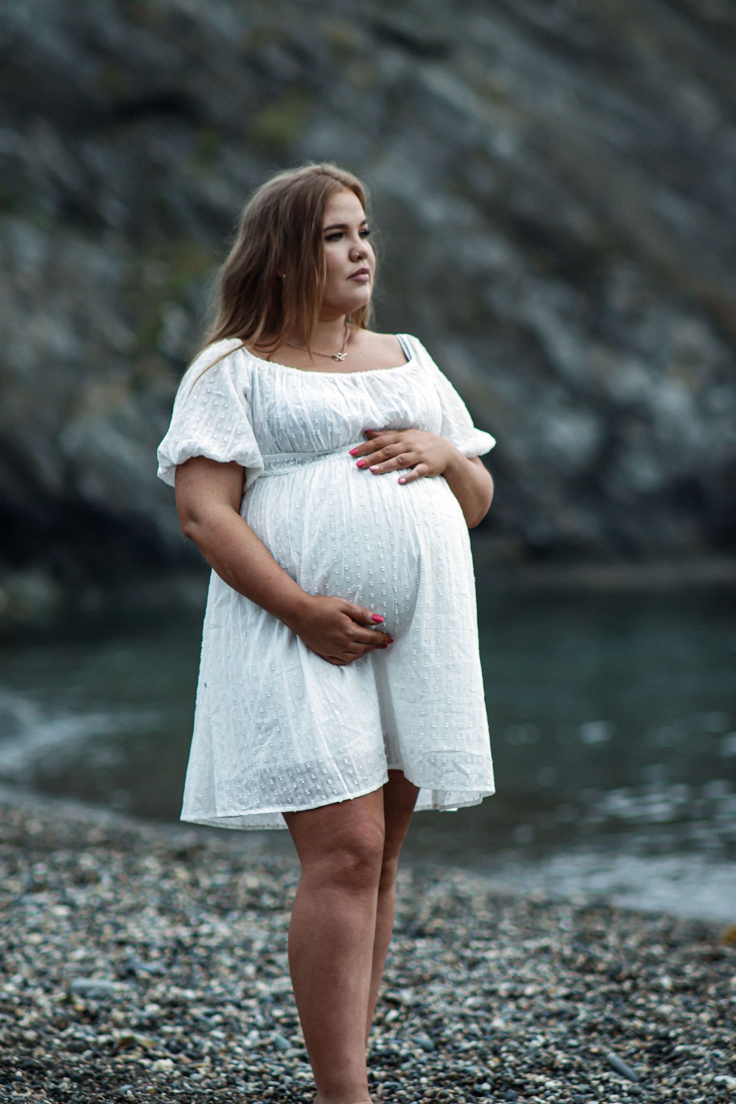 Woman in white dress poses on rocky beach shore against dramatic cliffs and water.