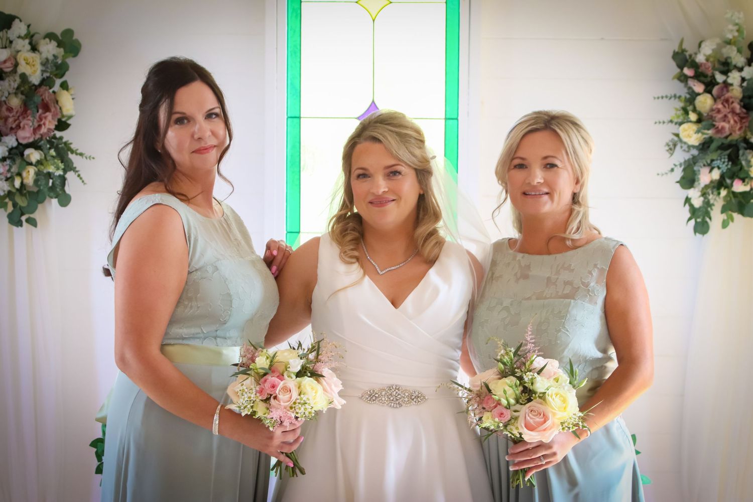 Bridesmaids in soft blue dresses pose with bouquets near stained glass window.