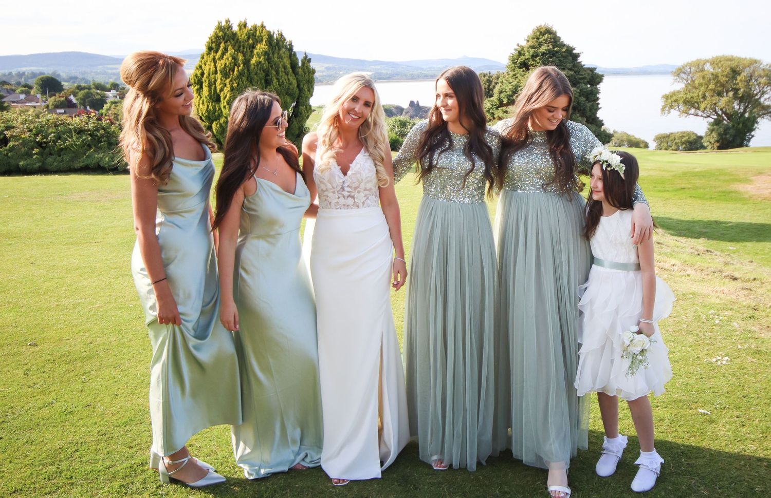 Group of bridesmaids in pastel dresses standing together on a sunny lawn by the water.