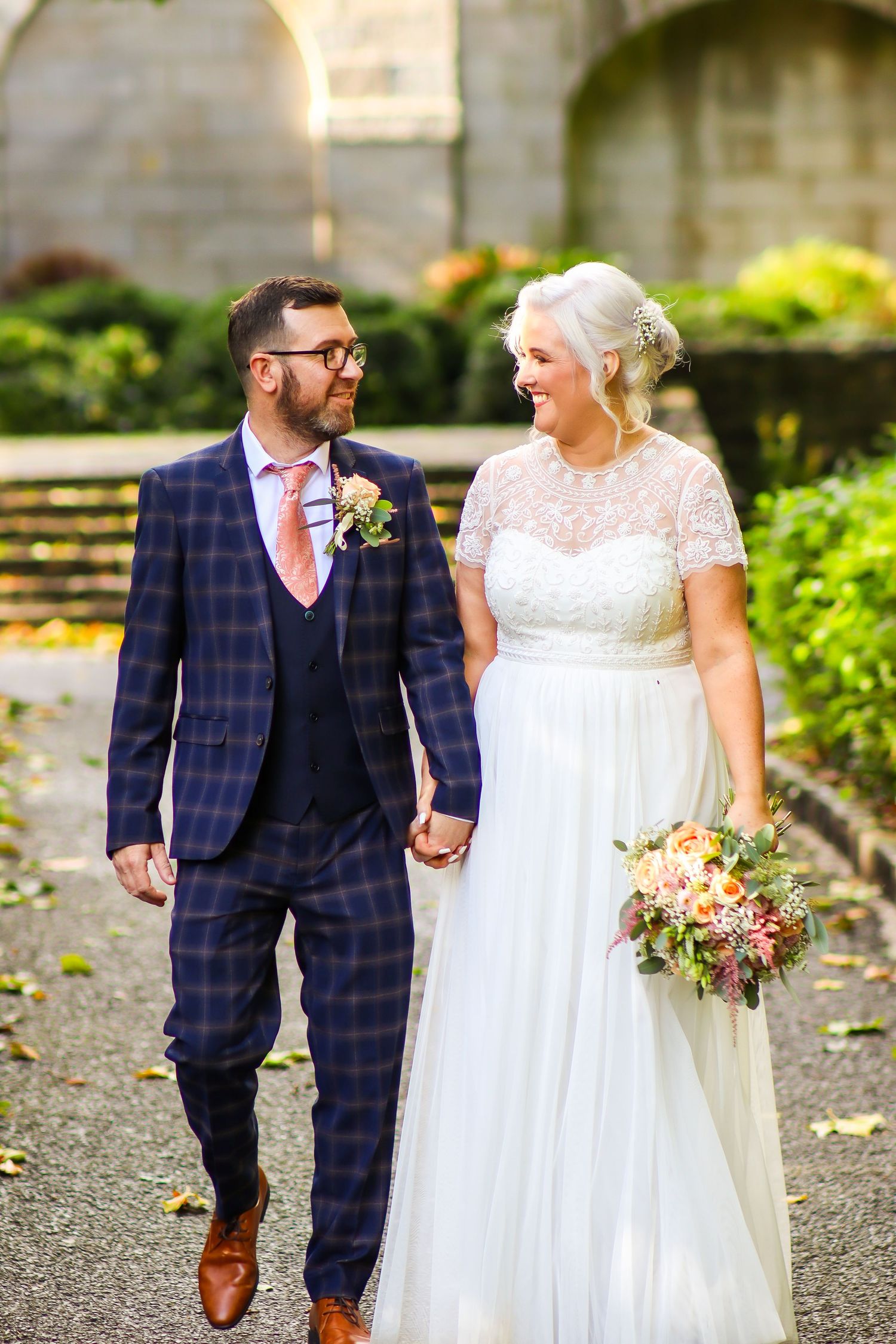 Newlyweds walk hand in hand outdoors, bride in lace dress and groom in plaid suit.