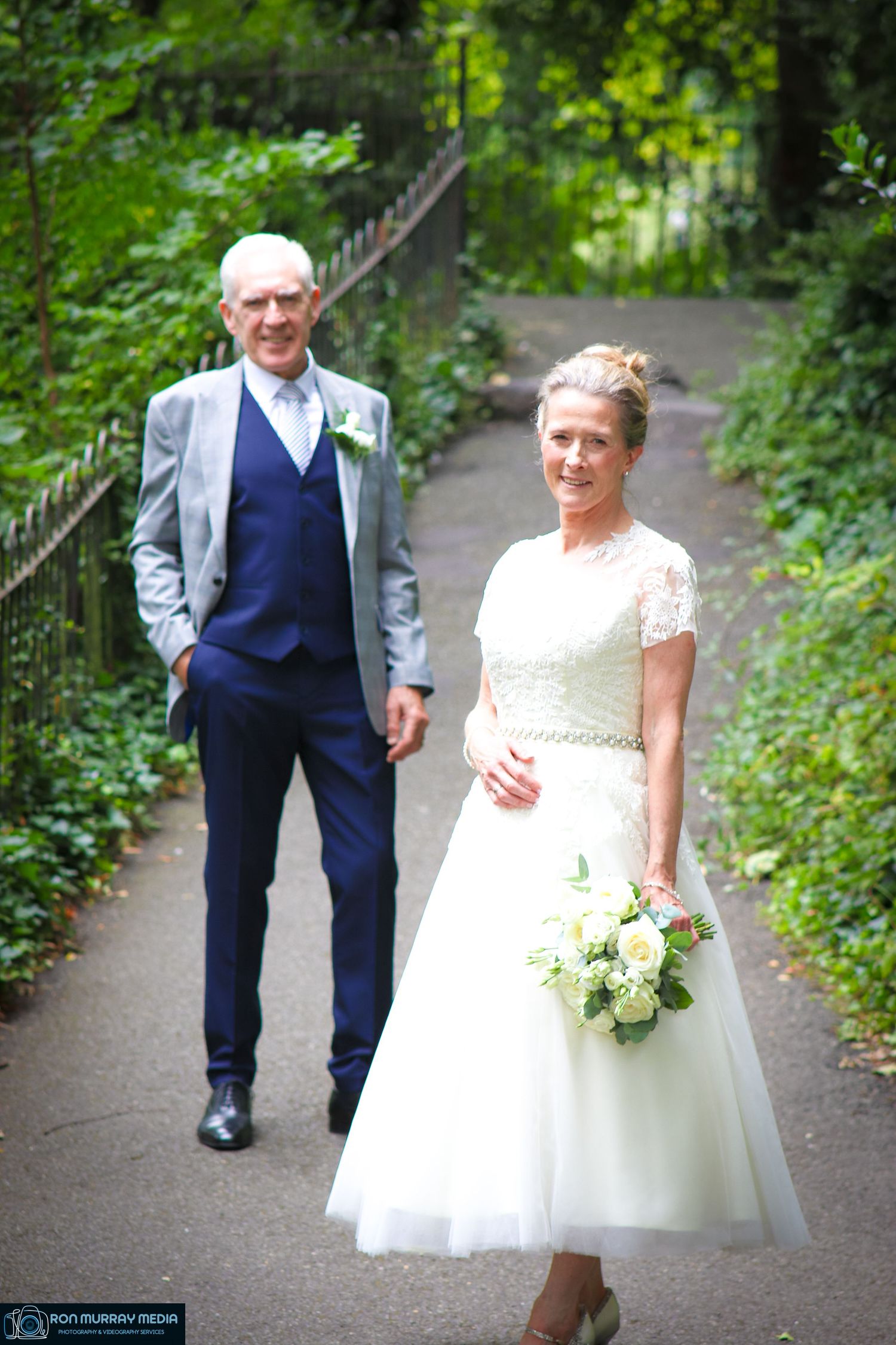 A wedding couple poses on a garden path surrounded by lush greenery wearing a tea-length dress and navy suit.
