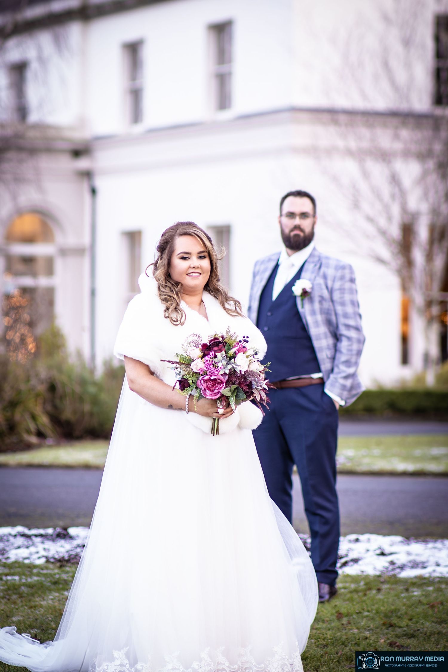 A winter wedding portrait in front of a white mansion with the bride holding a vibrant purple bouquet.