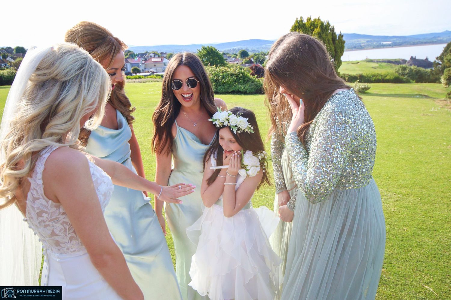 A group in light blue and white dresses laughs together on a sunny grassy lawn with mountains in the background.
