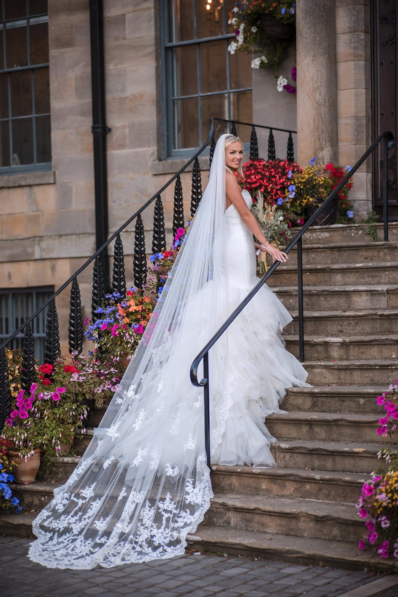 A bride in a flowing white gown with lace train poses on stone steps adorned with colorful flowers.