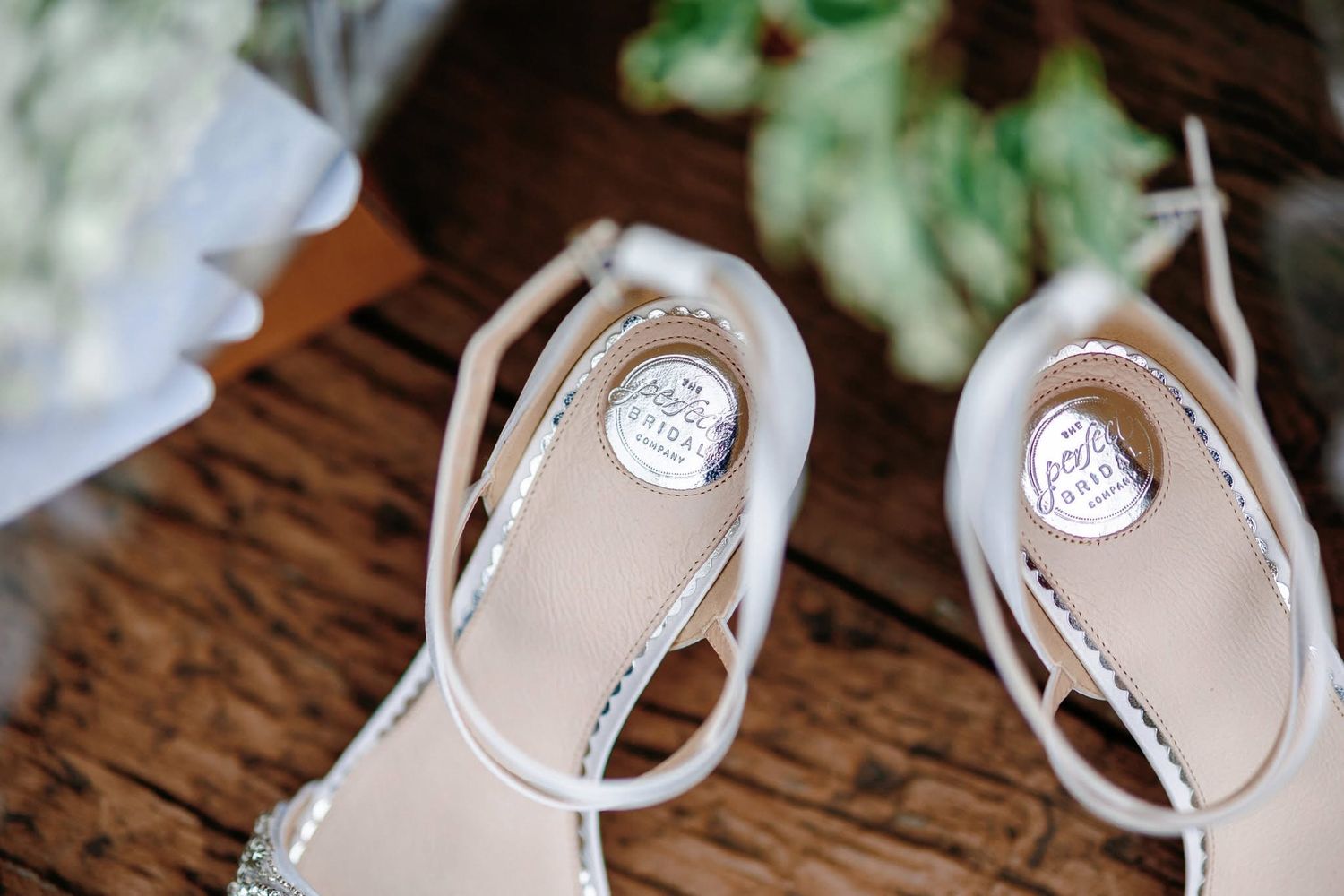 A pair of elegant white wedding shoes with delicate lace details sits on a rustic wooden surface.