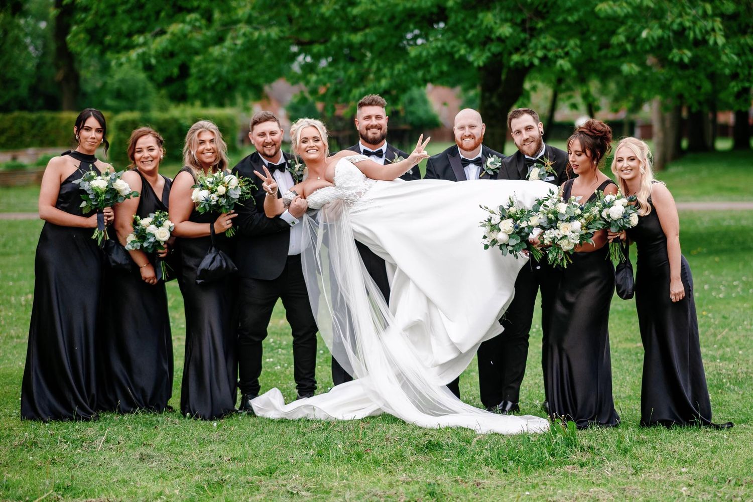 Wedding party in black dresses poses playfully with the bride's flowing veil outdoors.