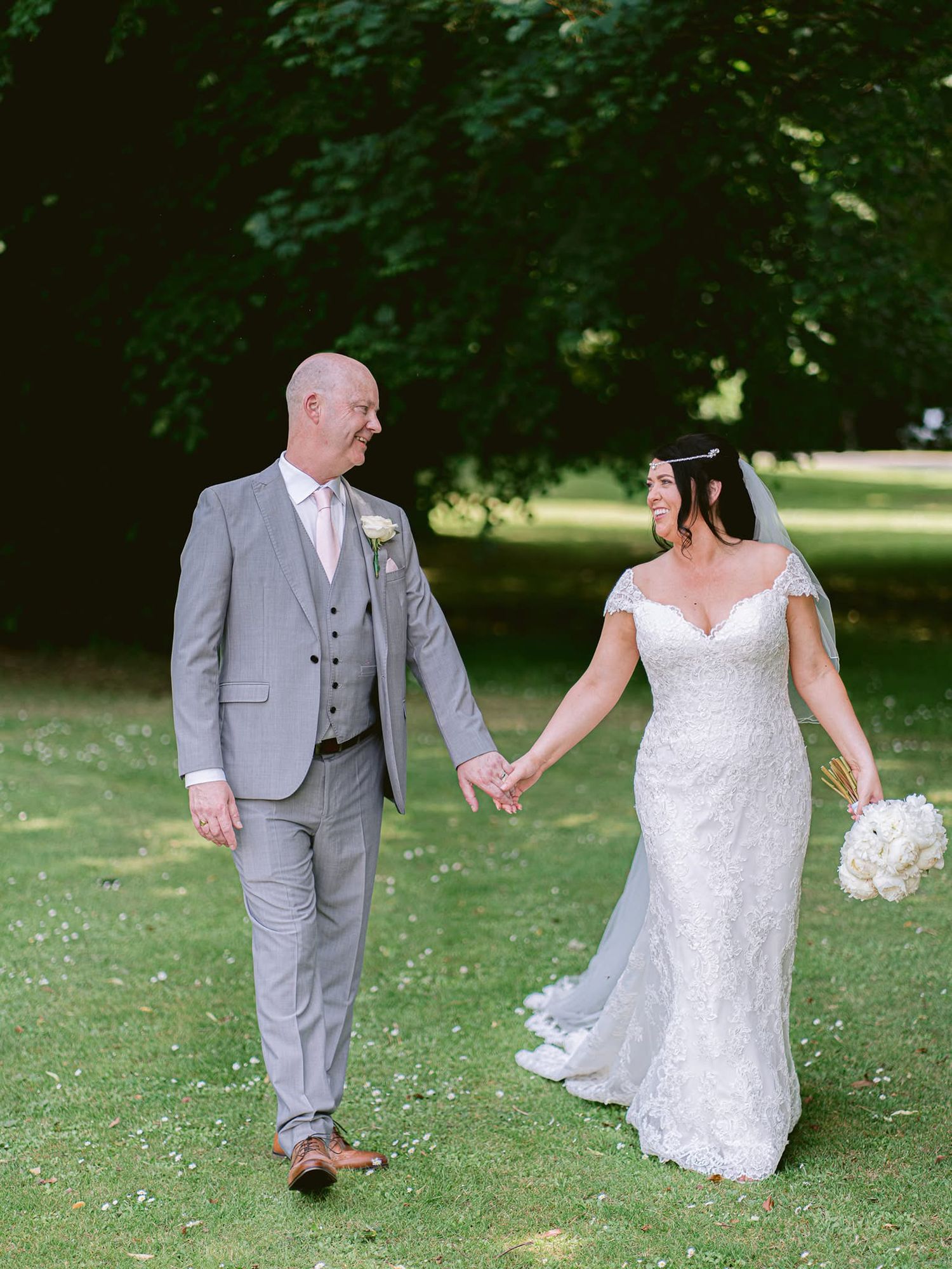 A couple in wedding attire dances playfully on a grassy lawn surrounded by trees.