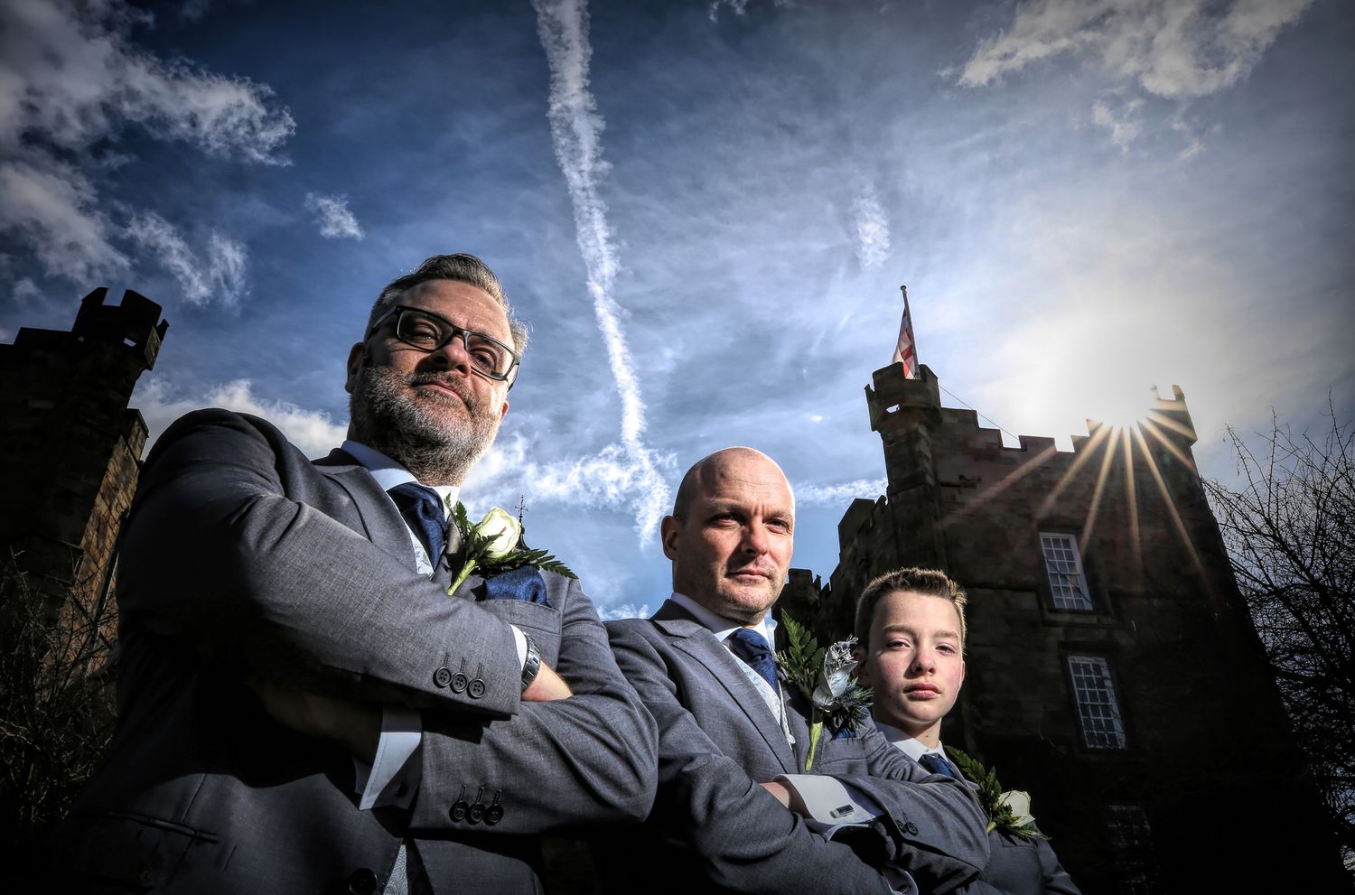 Three people pose dramatically against a dramatic sky with castle-like building in background.