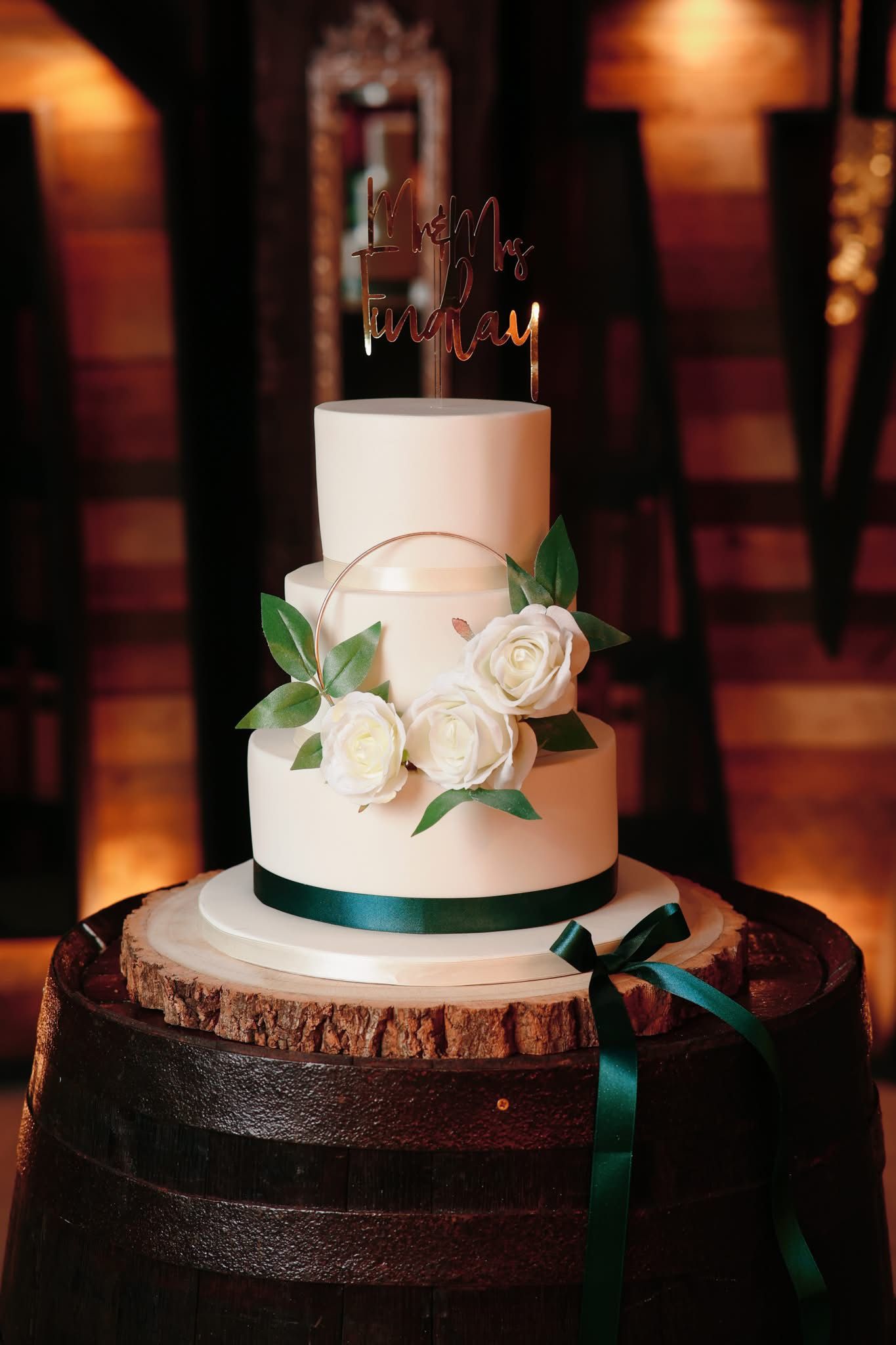 Elegant white wedding cake decorated with sugar roses and emerald green ribbons on a rustic wood stand.