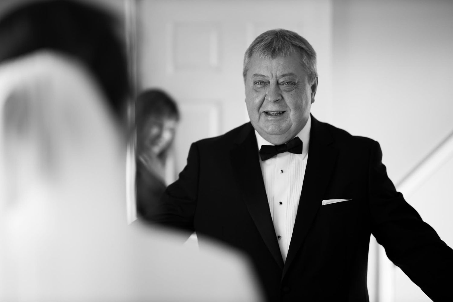 A person in a black tuxedo and bowtie gestures expressively while speaking at a formal event.