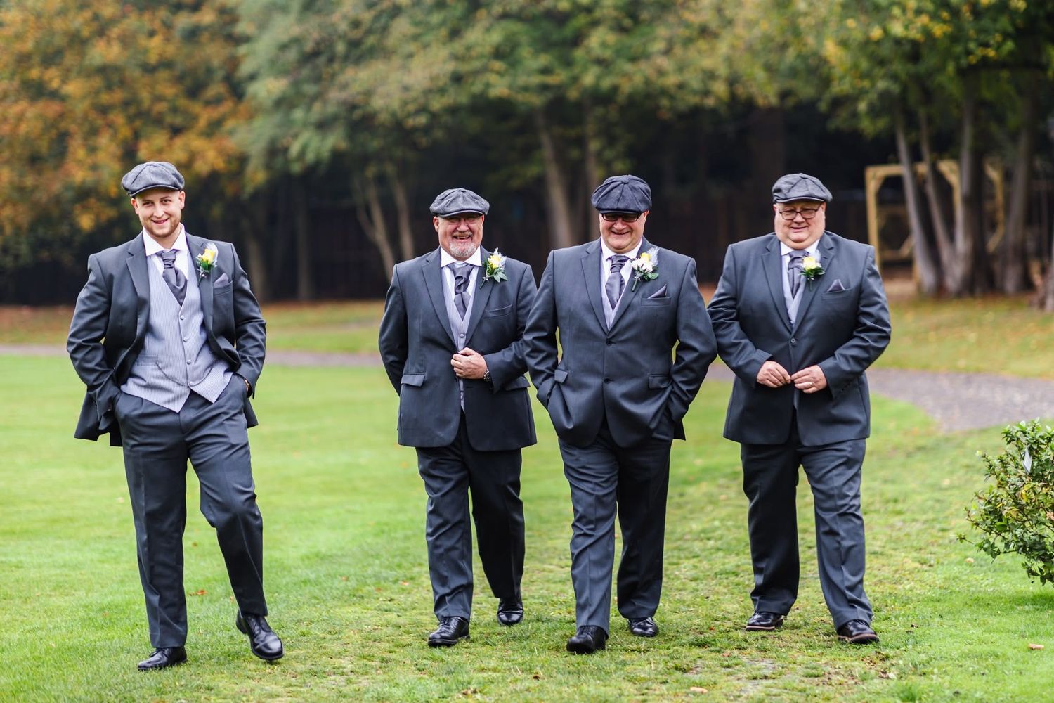 A group of groomsmen in matching navy suits perform a synchronized jumping photo on a green lawn.