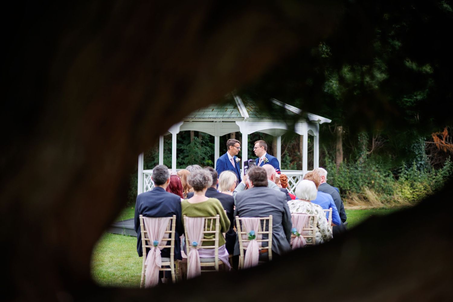 Guests seated in white chairs during an outdoor wedding ceremony at a white gazebo in a garden setting.