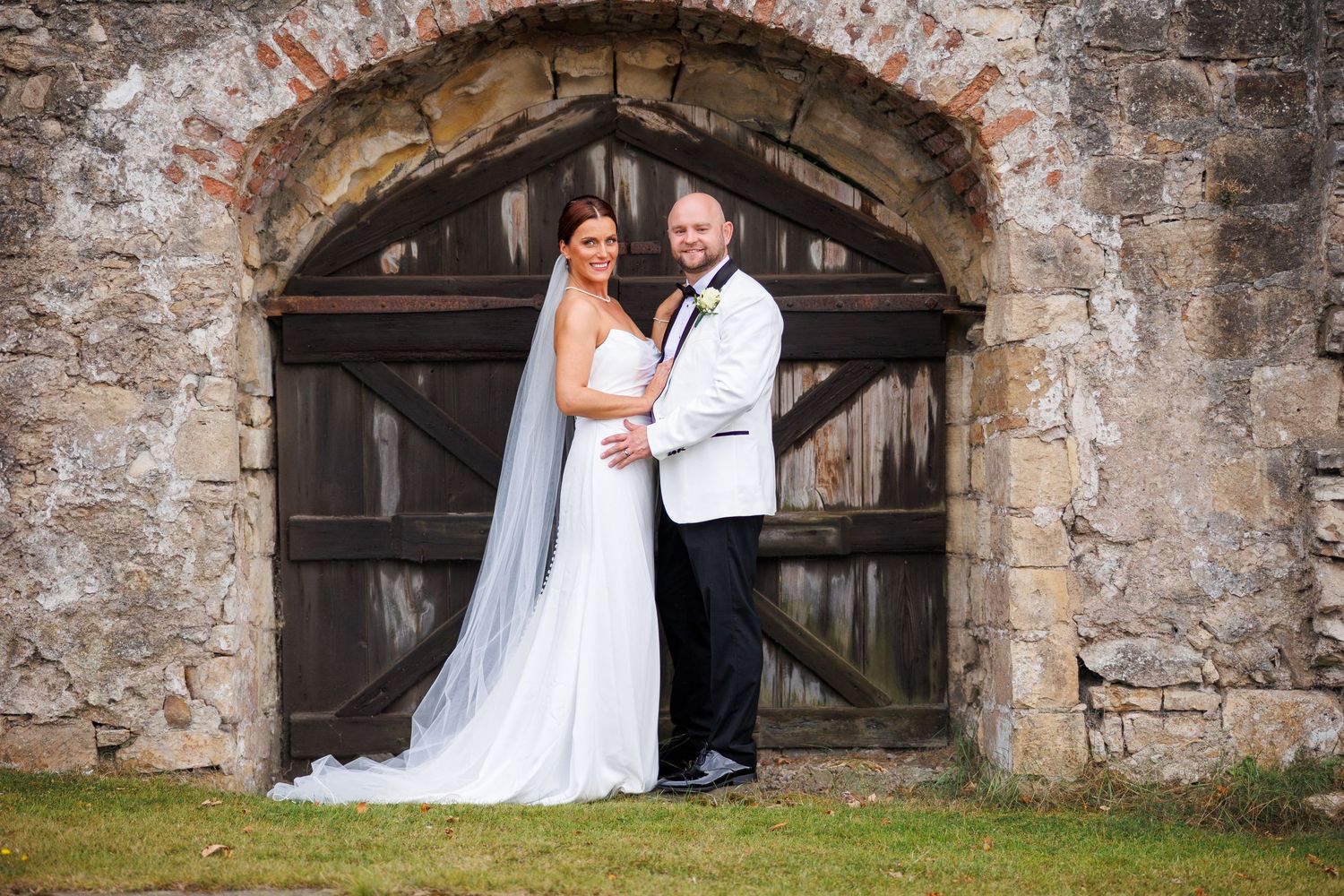 A newlywed couple poses in front of a rustic stone archway with wooden doors at their wedding.