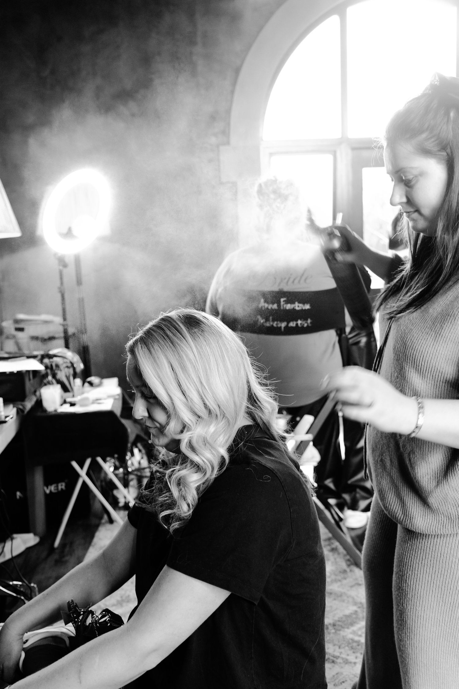 A black and white photo of a stylist working on a client's hair in a salon with dramatic lighting and smoke effects.