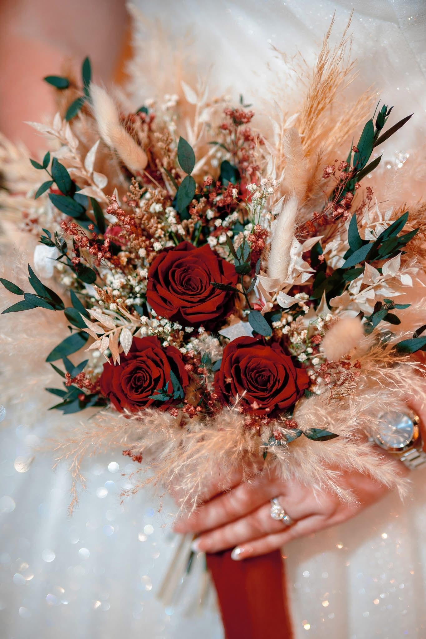 A romantic floral arrangement featuring deep red roses, dried pampas grass, and delicate white baby's breath on a soft pink background.