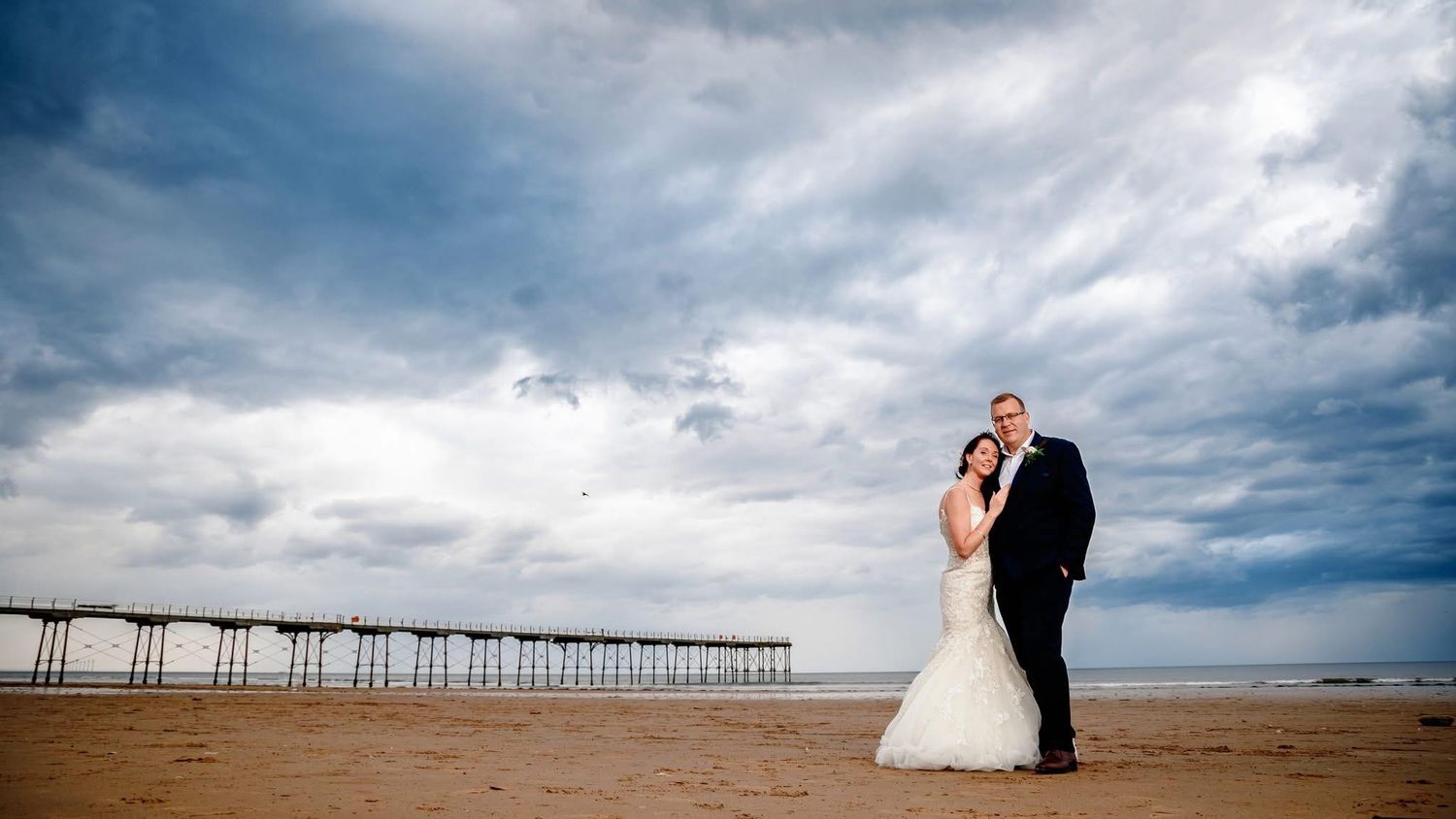 Wedding couple embraces on beach with dramatic pier and cloudy sky in background.