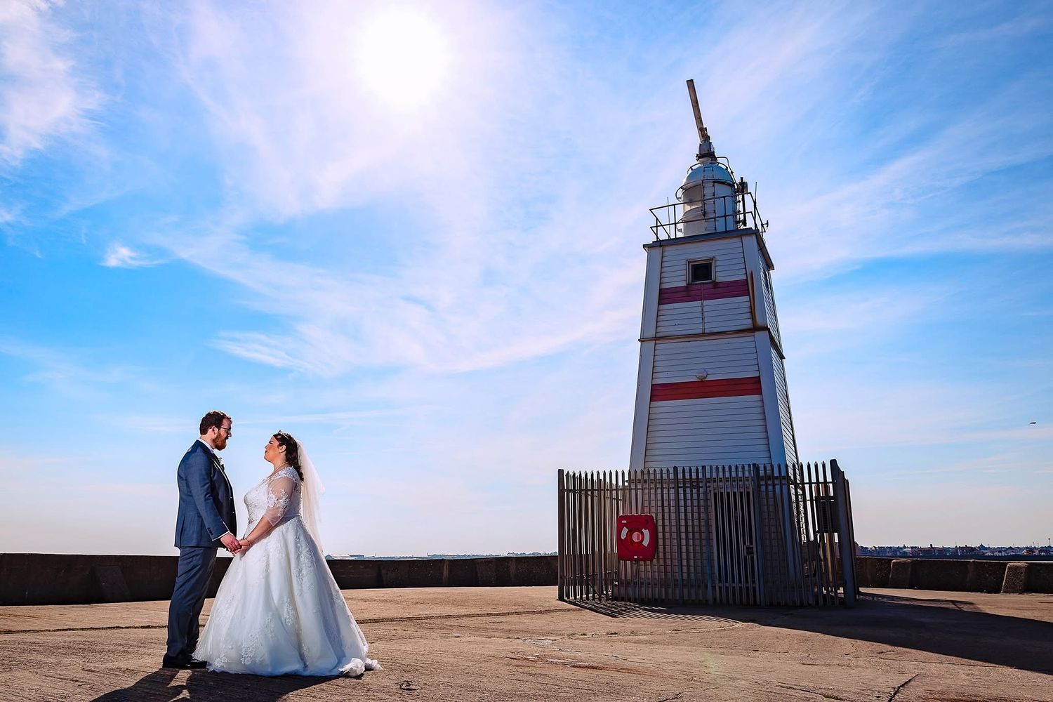 A lighthouse stands tall against a blue sky while a couple shares a romantic moment on the beach.