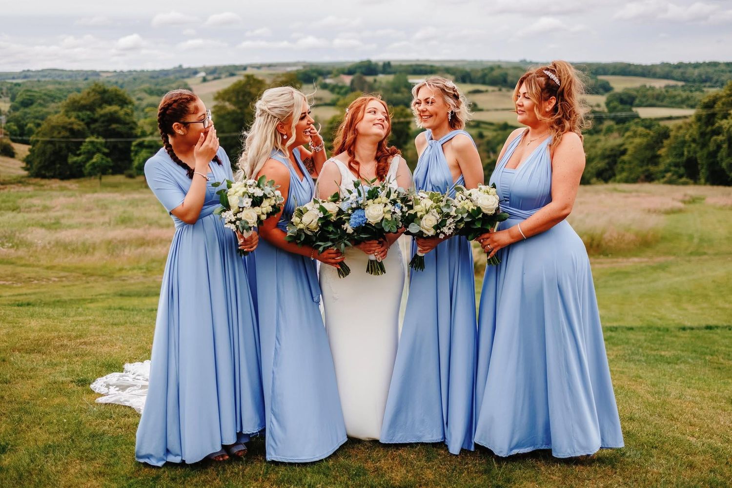Bridesmaids in light blue dresses pose together on a hilltop with bouquets.