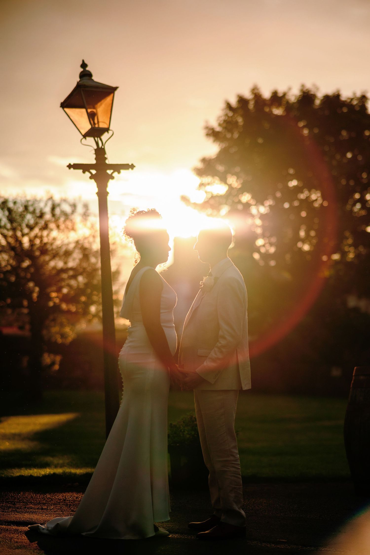 A romantic silhouette of a couple standing by a vintage lamppost at sunset with golden light streaming through trees.
