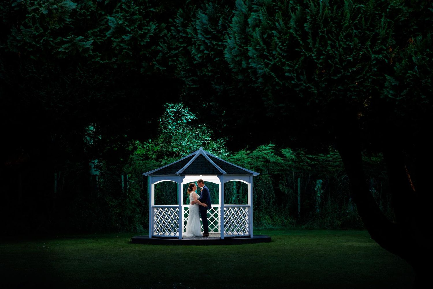 Illuminated white gazebo glows in dark garden setting.