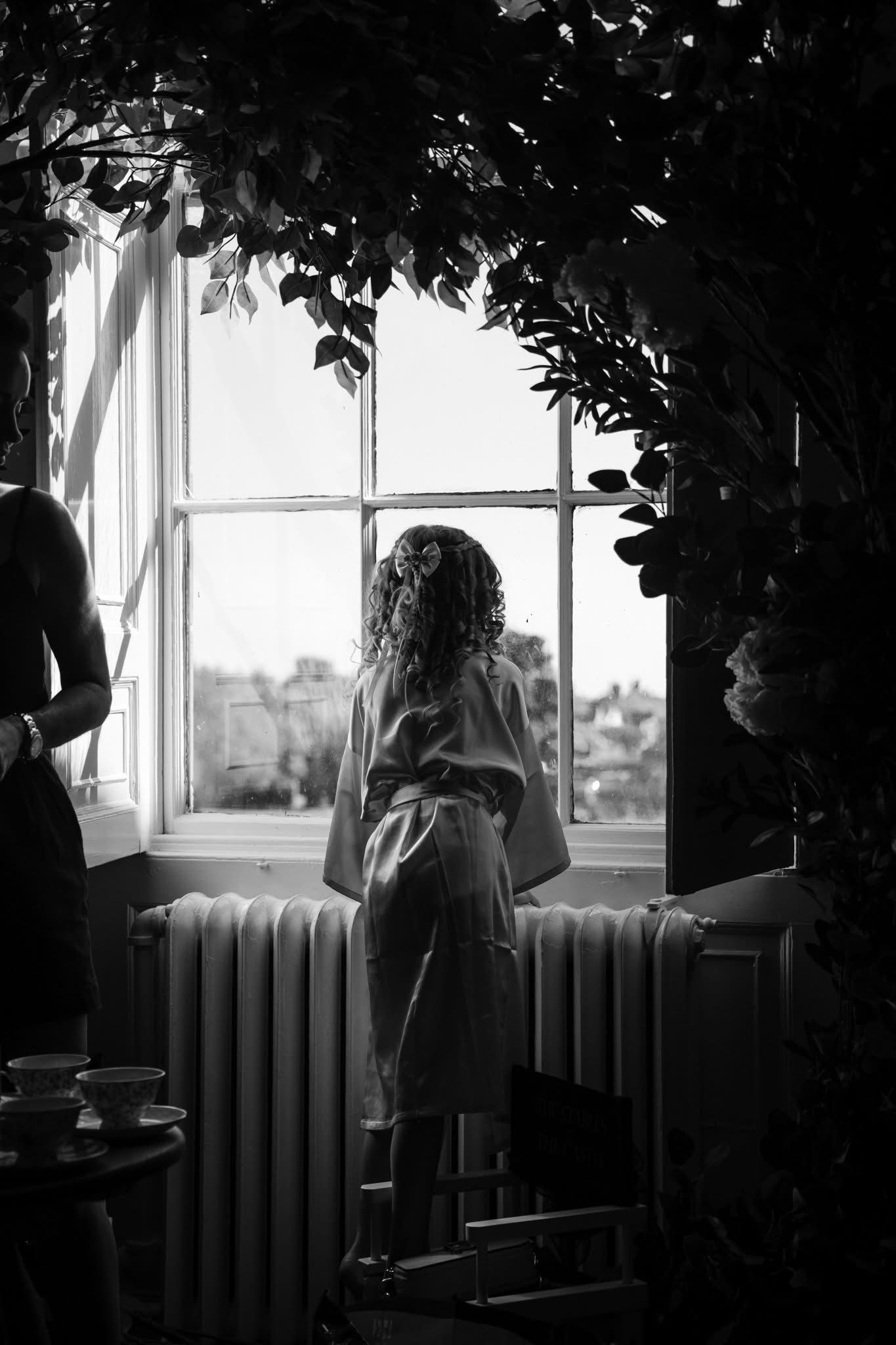 A silhouetted figure stands by a window with radiator and foliage framing the moody black and white scene.