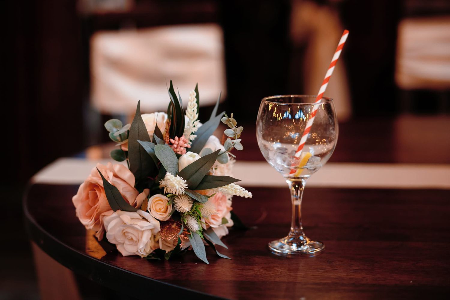 A romantic arrangement of roses and eucalyptus sits beside a wine glass with straw on a dark wooden table.