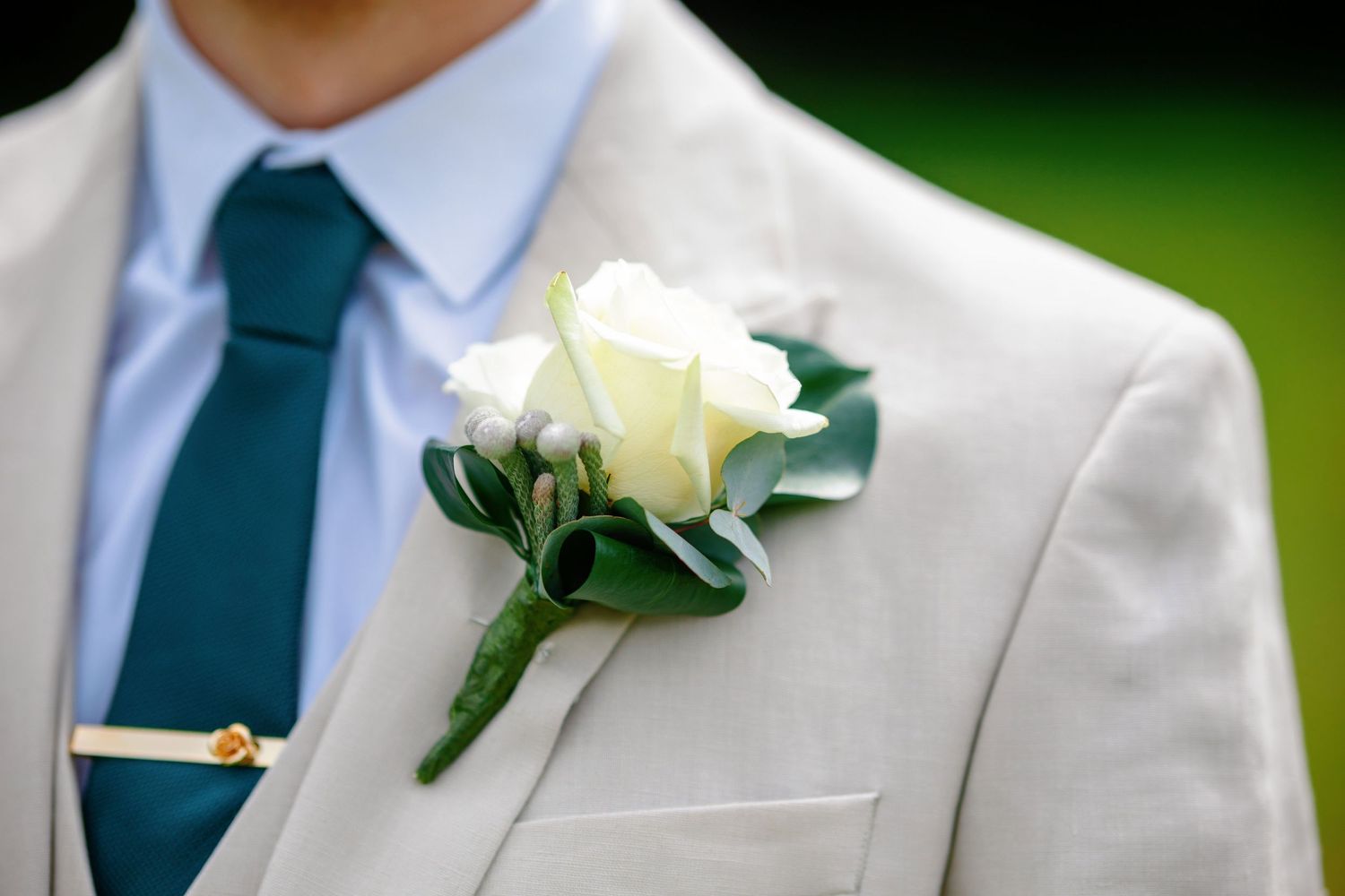 Close-up detail of a white rose boutonniere pinned to a light gray suit jacket with emerald green tie.