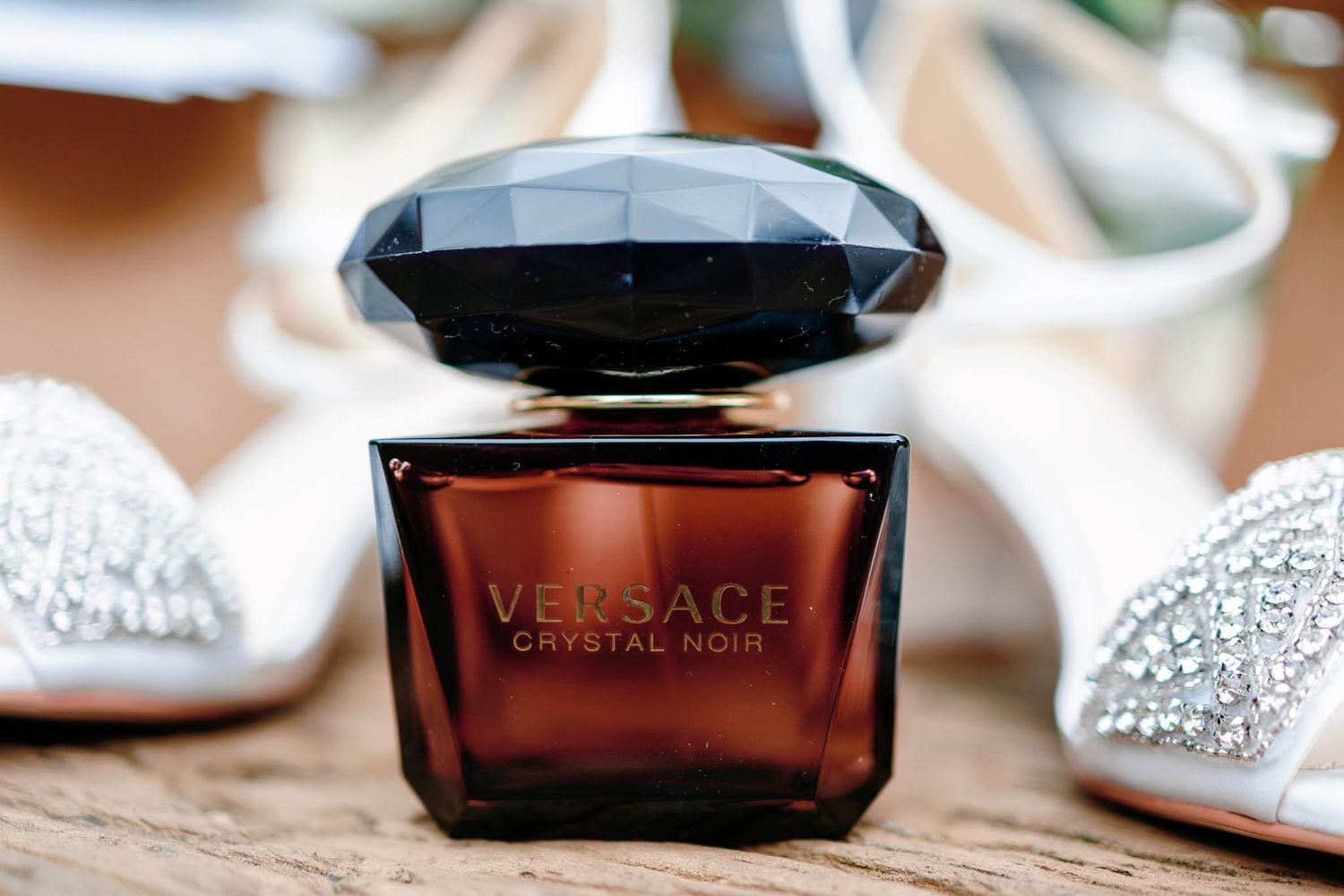 A bottle of Versace Crystal Noir perfume sits elegantly between sparkling silver shoes on a wooden surface.