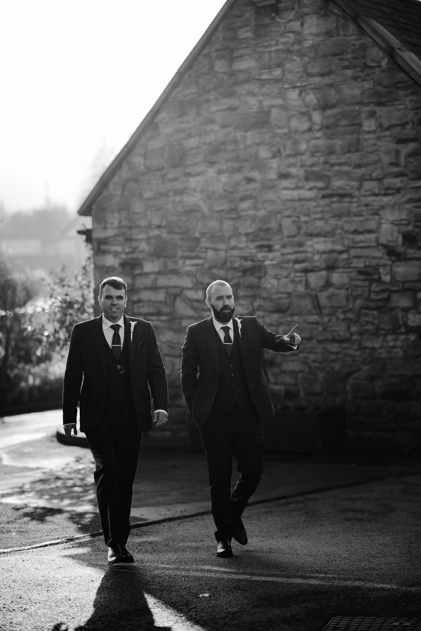 Two figures in dark suits walk together on a stone path in dramatic black and white lighting.