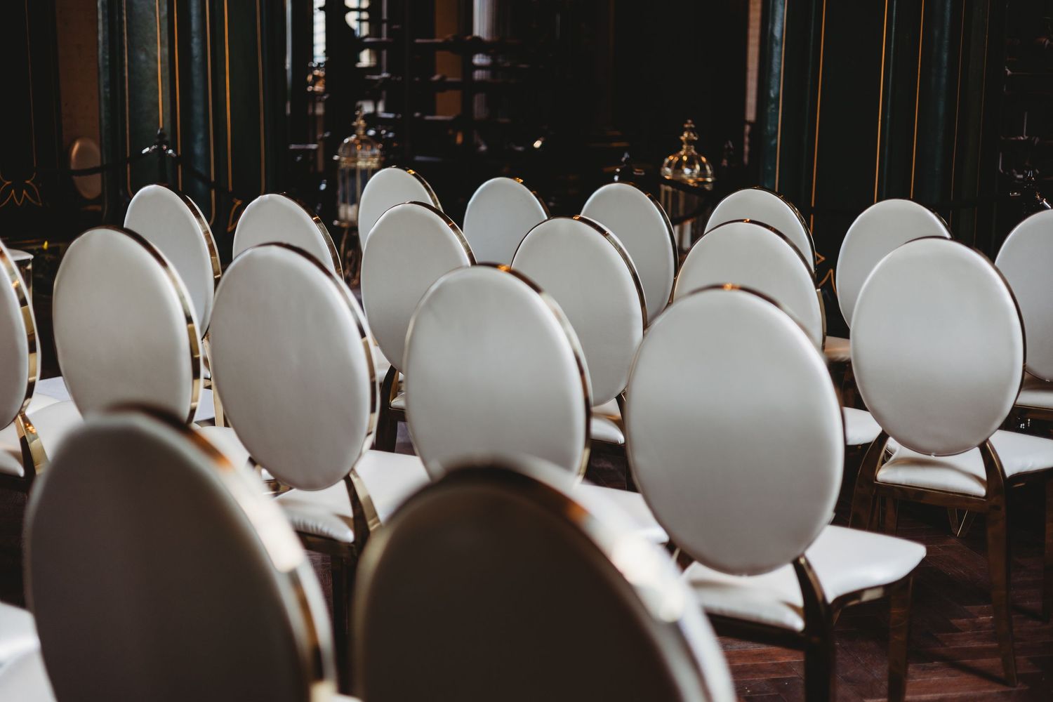 Empty white chairs arranged in rows at an indoor venue with dark lighting and wooden floors.