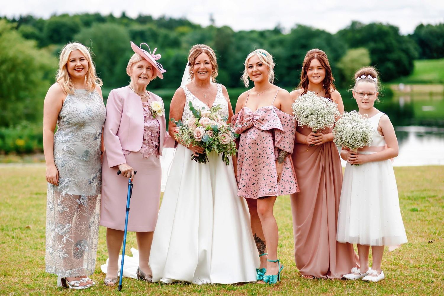 Bridal party in pastel dresses poses together by a lake on a sunny day.