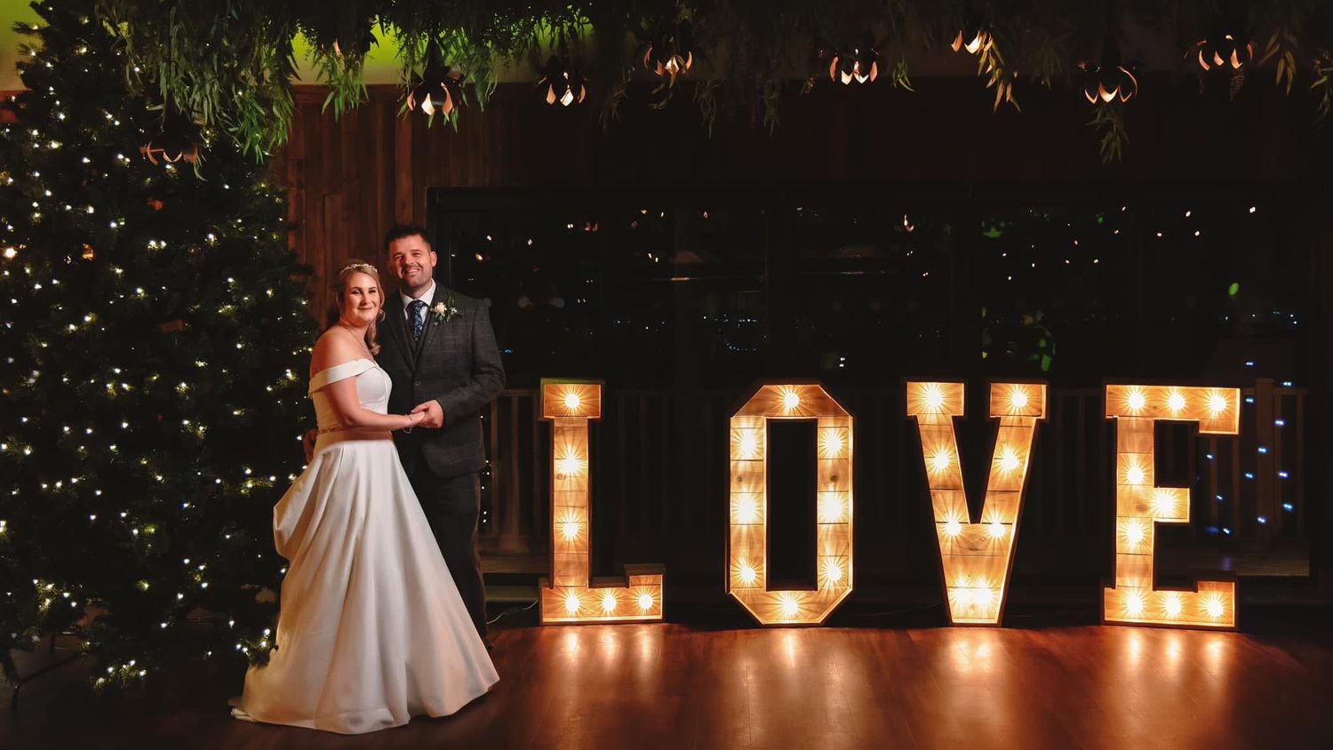 A romantic outdoor wedding venue at night with illuminated LOVE marquee letters glowing against dark foliage.