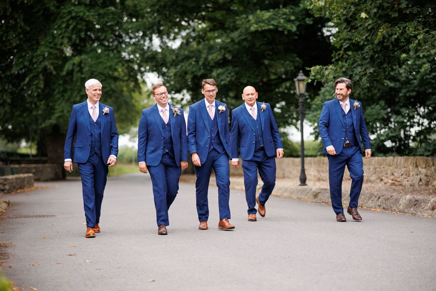 Groomsmen wearing matching navy blue suits walk together on a tree-lined path during a wedding celebration.