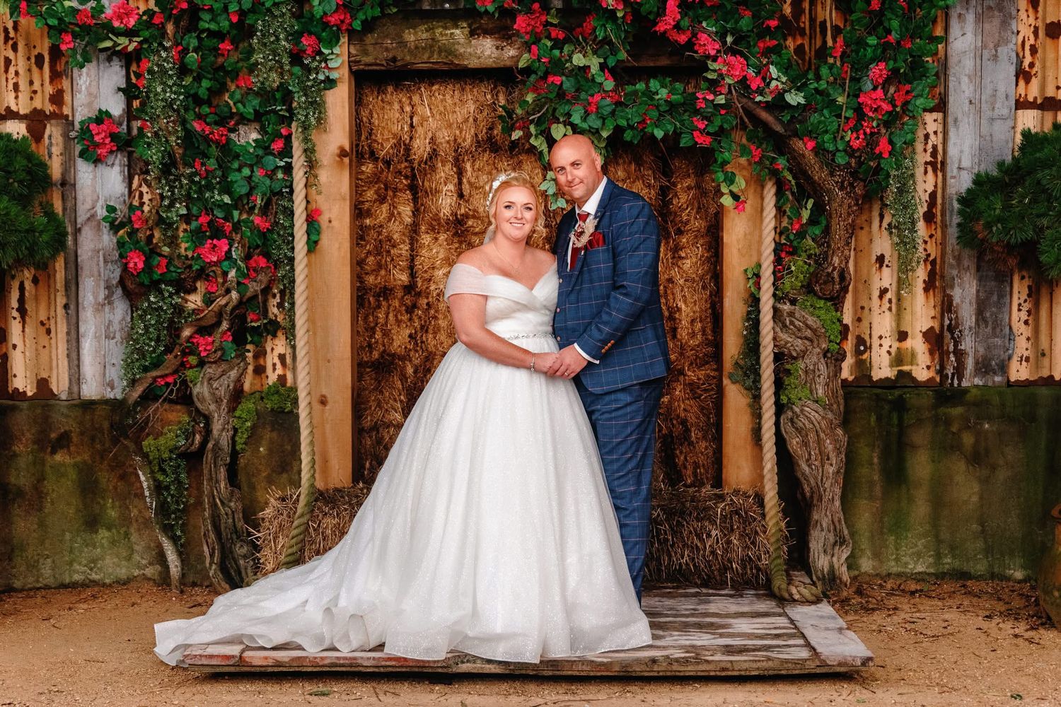 A romantic wedding couple poses beneath a rustic wooden arch adorned with climbing red roses in a garden setting.