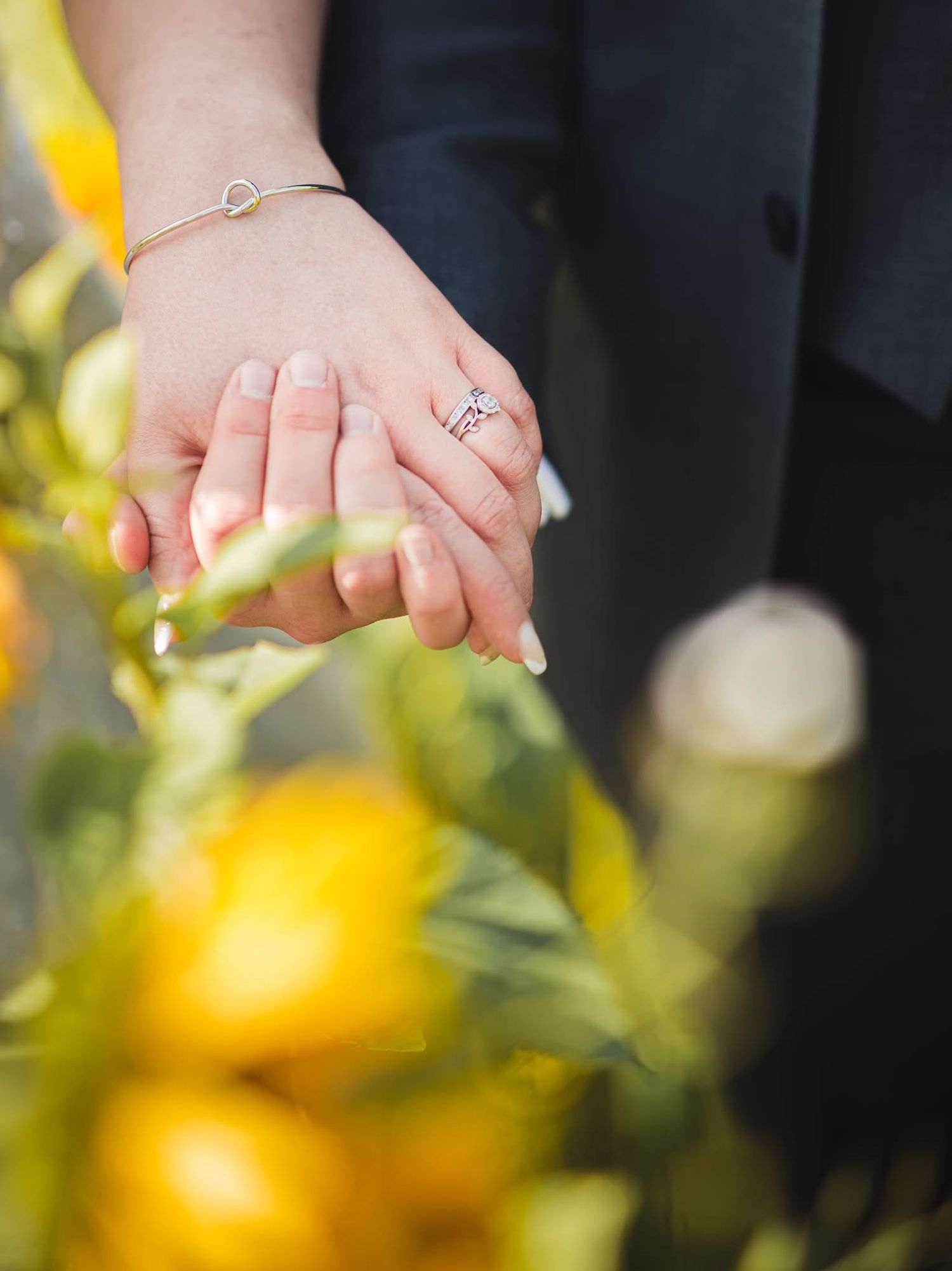 Close-up of hands with engagement rings intertwined against a blurred yellow and green floral background.