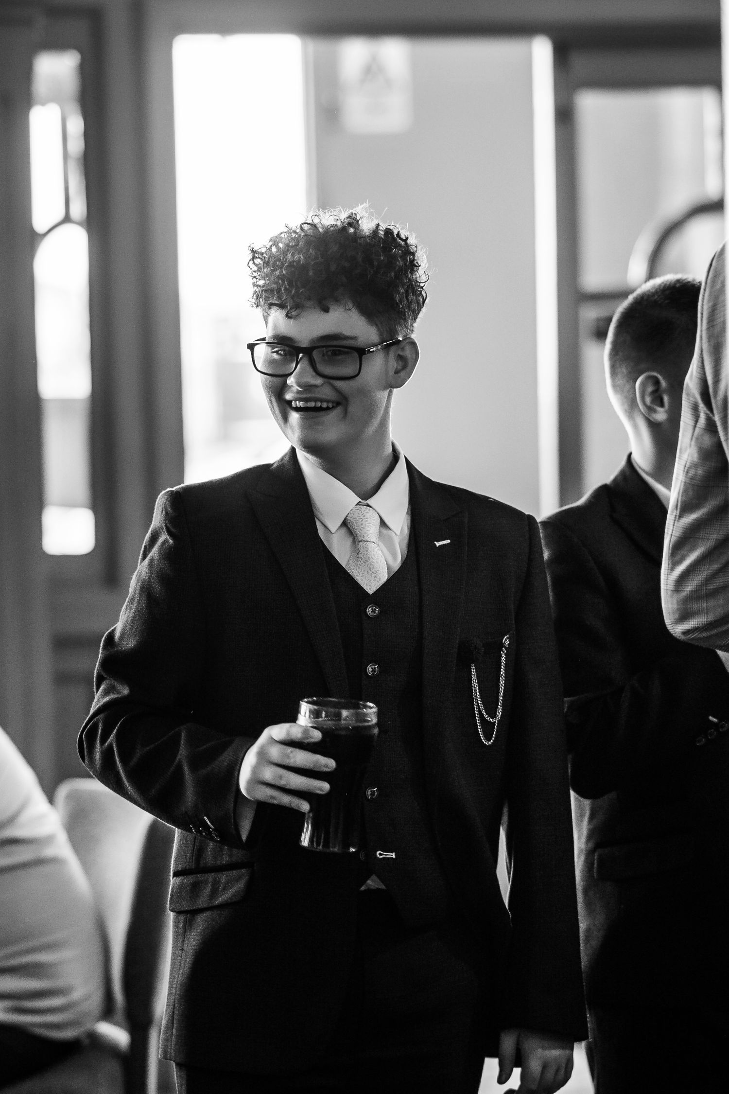 A person in a dark suit and glasses holds a drink at an indoor event in black and white photography.