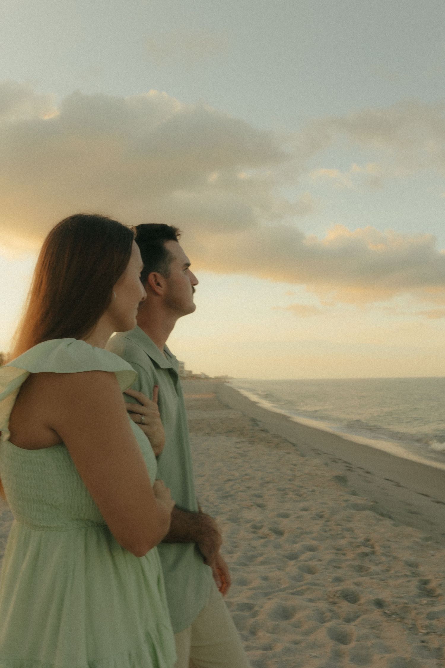 A couple in mint green outfits embraces while watching a beautiful sunset on the beach.
