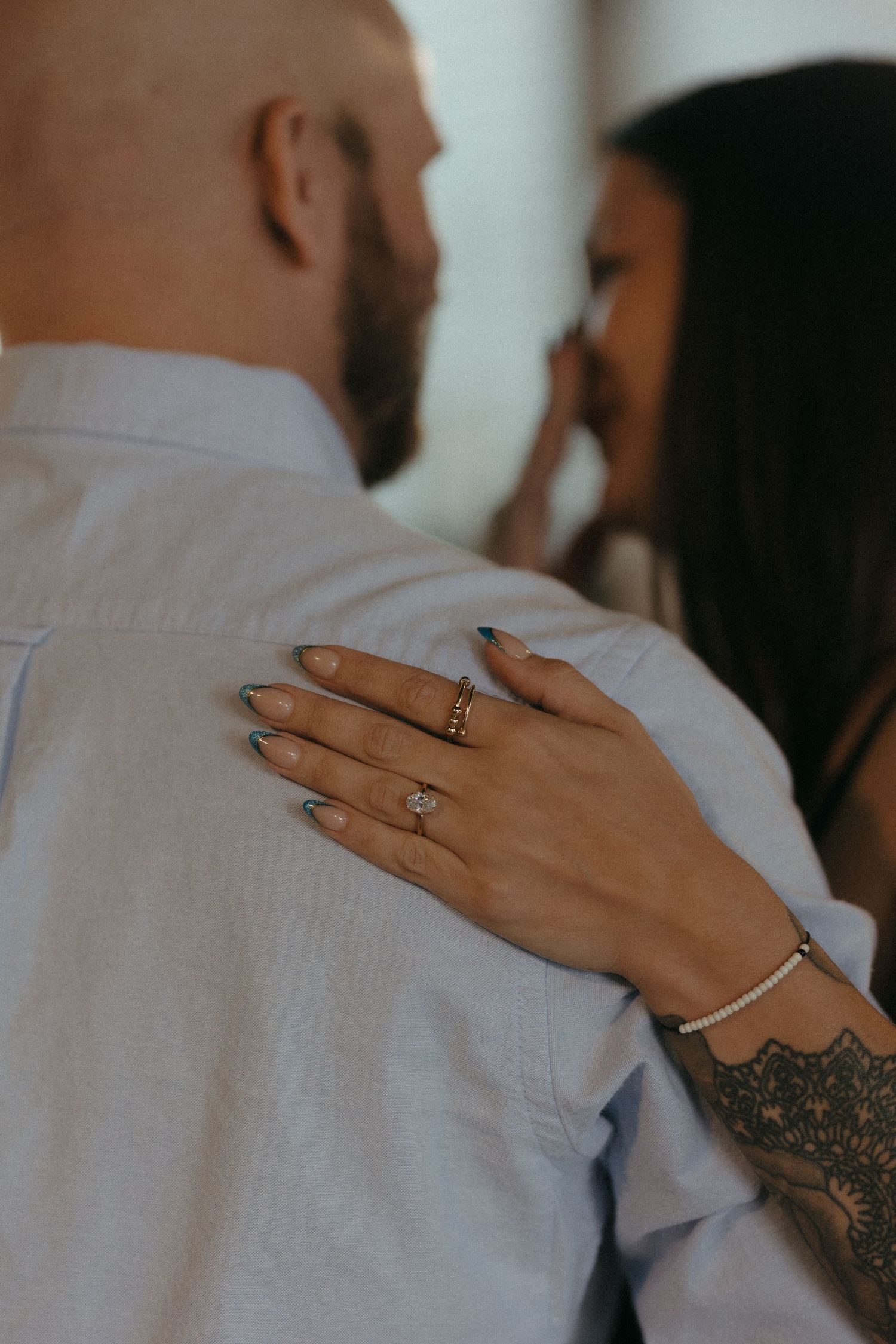 Close-up of a woman's tattooed hand with rings resting on a man's shoulder as they embrace.