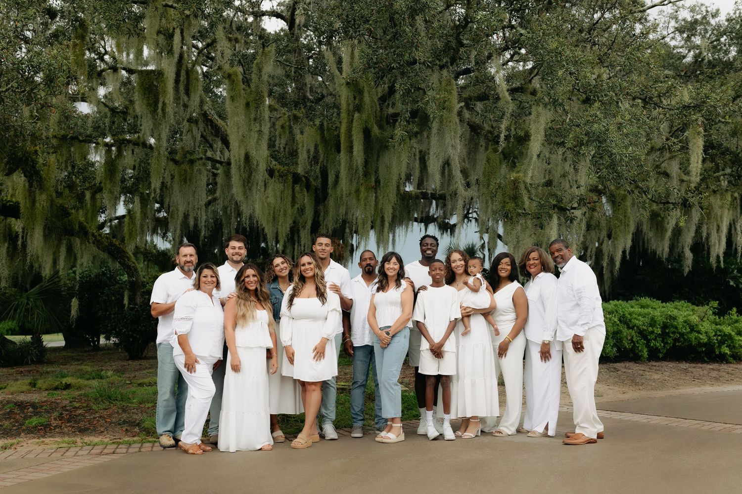 Large family group portrait in white outfits under Spanish moss-draped oak trees outdoors.