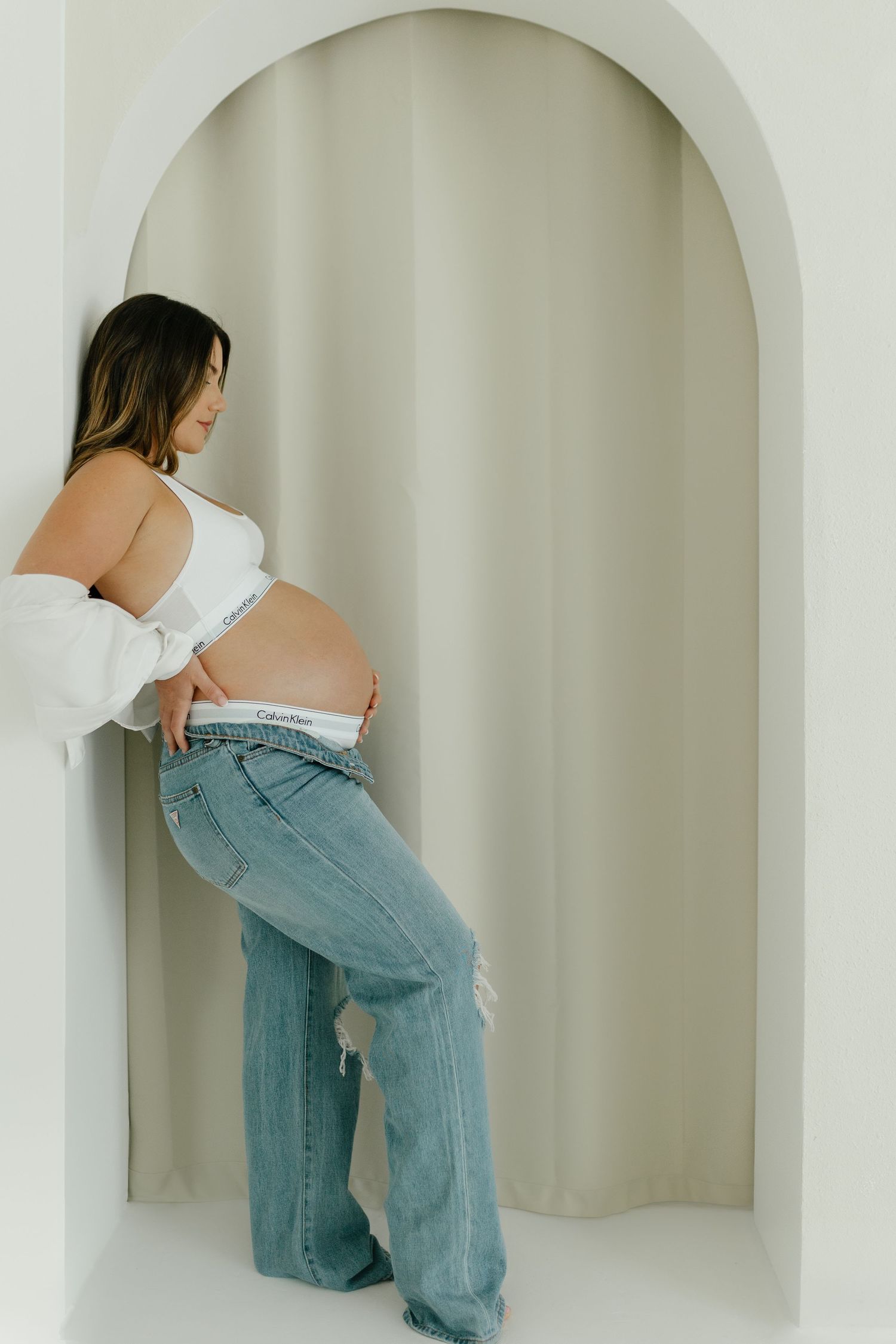 Pregnant woman in white top and jeans leaning against an arched wall holding her baby bump.