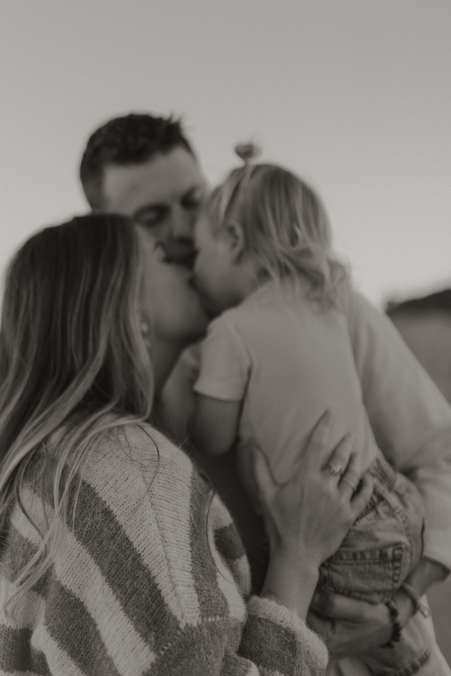 Black and white photo of a couple kissing while holding their young toddler child in an outdoor setting.