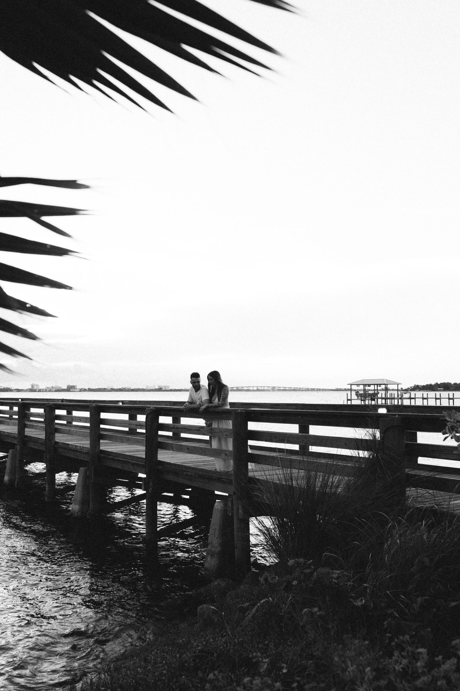 Black and white silhouette of couple on a waterfront bridge framed by palm fronds.