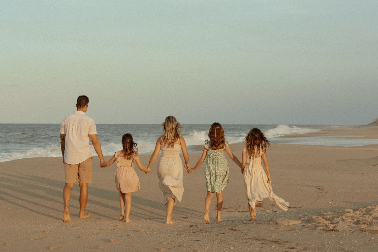 Family of five walking hand-in-hand along a sandy beach at sunset, viewed from behind.
