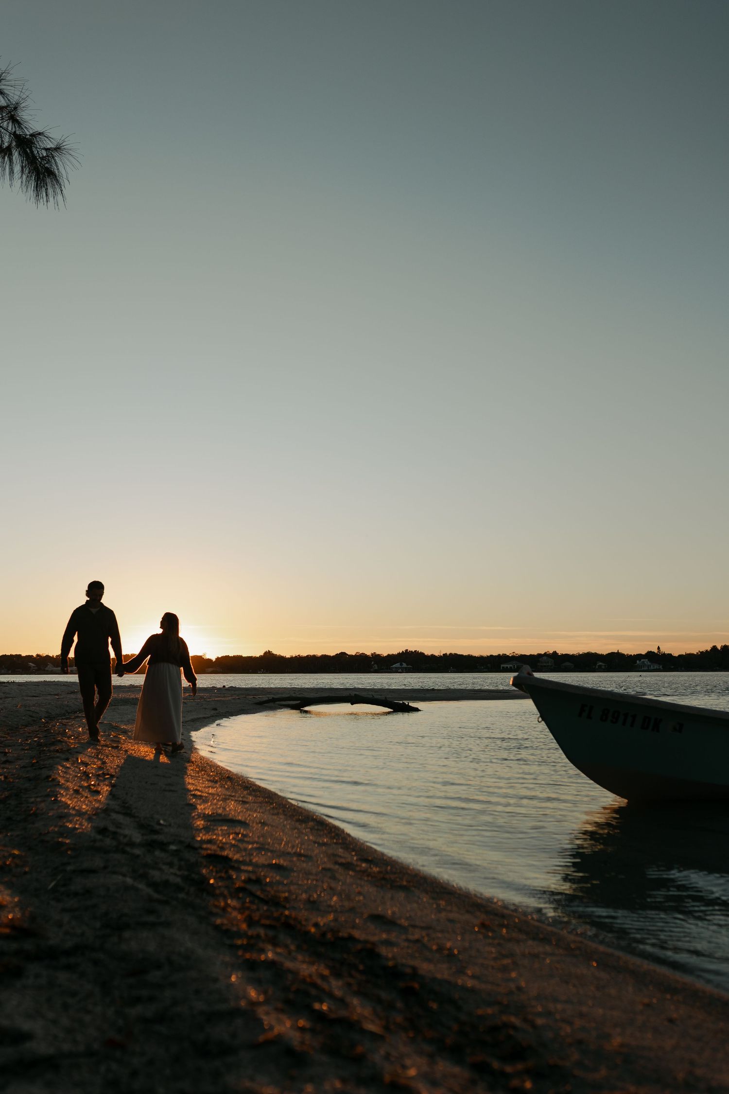Couple holding hands walking along sandy beach at sunset with boat and calm water in background.